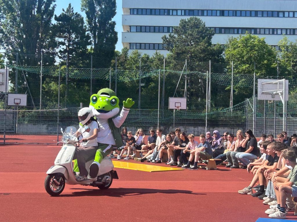 Groene mascotte op een scooter Hoogwaardig mascotte pak – een groene mascotte op een scooter rijdt voor een groep toeschouwers op een sportveld tijdens een evenement. Het comfortabele mascotte pak heeft realistische details en is ontworpen voor optimale bewegingsvrijheid en ventilatie.